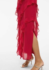 Raudona šifoninė suknelė su raukšlėmis ir šoniniu plyšiu, dėvima su sidabriniais strappy aukštakulniais, pabrėžianti pusiau permatomą tekstūrą ir laisvą dizainą.