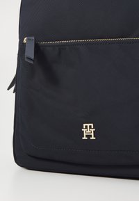 Navy fabric backpack with a textured surface, gold zipper, and a gold logo accent. Features a front pocket with a zip closure.