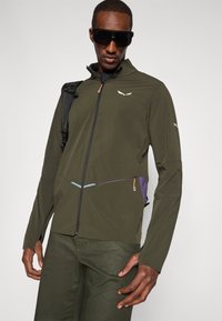 Ελιά πράσινο μπουφάν με φερμουάρ, σκούρα αξεσουάρ, μια διακριτική λογότυπο και λεπτομέρειες με μπλοκάρισμα χρωμάτων. Διαθέτει υφαντό ύφασμα και πλαϊνές τσέπες.