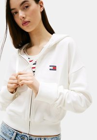 Felpa bianca con zip, dal tessuto morbido, con polsini e orlo a coste, e un piccolo badge con il logo "Tommy Jeans" sul petto.