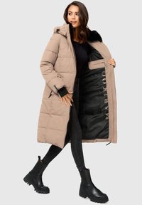 Abrigo acolchado beige con capucha, diseño acolchado y forro negro con marca impresa. Combinado con vaqueros ajustados negros y botines negros.