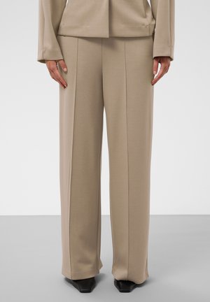 Personne portant un pantalon beige à jambes larges et un blazer assorti, debout les mains détendues le long du corps devant un fond gris et blanc.