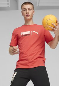 Rode katoenen t-shirt met witte "PUMA HANDBALL" tekst en logo, gecombineerd met zwarte broek. Het model houdt een gele handbal vast.