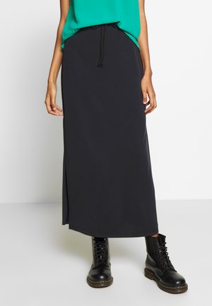 Jupe maxi noire avec fente sur le côté, fabriquée en tissu doux. Associée à un haut vert et des bottes noires à lacets. Design simple avec taille élastique.