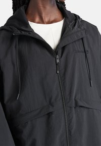 Giacca nera leggera con cappuccio, dotata di zip frontale, coulisse elastiche e tessuto con trama sottile. Logo del marchio sul petto.