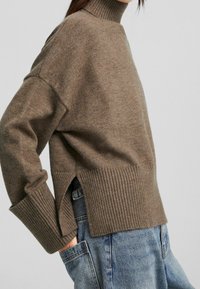 Brązowy sweter robiony na drutach z wysokim kołnierzem, ściągaczami na mankietach i bocznymi rozcięciami, w połączeniu z niebieskimi dżinsami. Miękka tekstura, luźny krój.