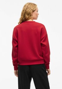 Sweatshirt rouge à manches longues, col rond, avec des poignets et un ourlet côtelés. Texture lisse, coupe ample et design minimaliste. Pantalon noir visible.