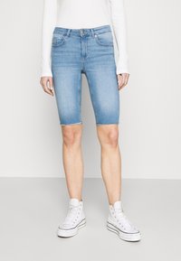 Bermudas de denim azul claro con dobladillo sin terminar, cinco bolsillos y una forma ajustada, combinadas con zapatillas altas blancas.