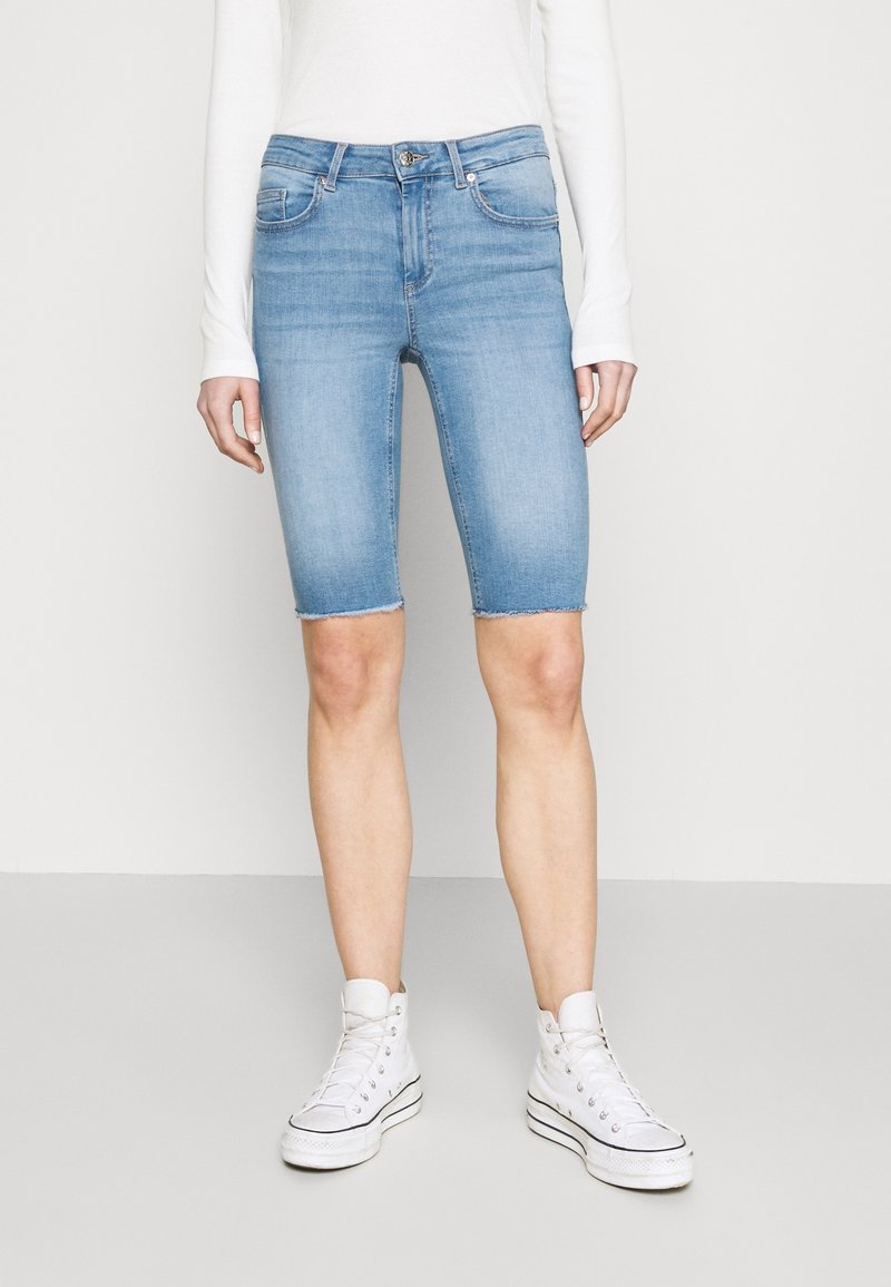 Bermudas de denim azul claro con dobladillo sin terminar, cinco bolsillos y una forma ajustada, combinadas con zapatillas altas blancas.