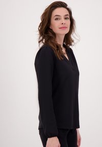 Vrouw met golvend bruin haar, gekleed in een zwarte blouse met lange mouwen en een zwarte broek, staand tegen een effen lichte achtergrond.