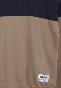 Beżowy sweter w poziome ciemnoniebieskie paski o miękkiej teksturze dzianiny, z logo Timberland na dole.
