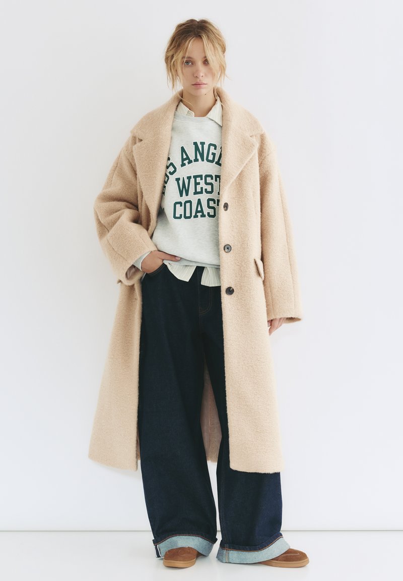 Beżowy, oversize'owy płaszcz z teksturowanej tkaniny, nałożony na szary sweatshirt z zielonym napisem, szerokie niebieskie dżinsy i brązowe buty.