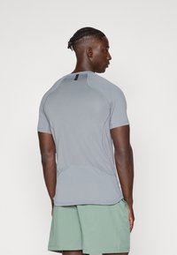Camiseta atlética gris de mangas cortas, con tejido texturizado, acentos de costura y un cuello alto. Se lleva con pantalones cortos de color verde claro.