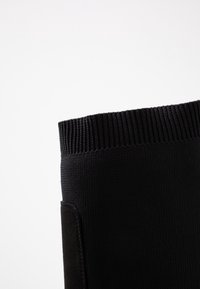 Chaussette noire avec une texture en tricot côtelé en haut et un tissu lisse à la base, dotée d'un petit détail de poche sur le côté.