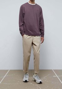 Fioletowa bluza z okrągłym dekoltem i ściągaczami, w połączeniu z beżowymi spodniami i białymi trampkami. Minimalistyczny design, bez widocznych wzorów.