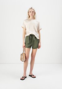 Lys rosa satin T-skjorte, løs passform med rullede ermer; olivengrønne shorts med snøring; svarte flip-flops; liten beige håndveske med flettet håndtak.