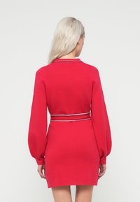 Czerwona dzianinowa sukienka z długimi bufkami, kołnierzykiem i wcięciem w talii z kontrastowym paskiem. Gładka tekstura, długość do kolan.