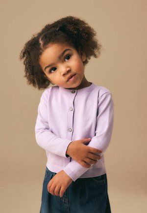 Fată tânără cu păr creț, purtând un cardigan lavandă și o fustă din denim, înclinând ușor capul cu brațele încrucișate în față.