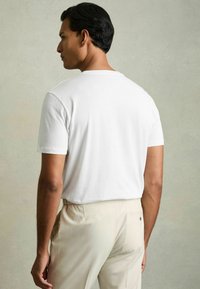 Tricou alb din bumbac, cu guler rotund, mâneci scurte și o croială lejeră, asortat cu pantaloni bej deschis, ce au un buzunar la spate.