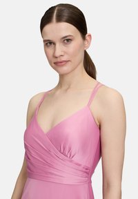 Różowa sukienka z cienkimi ramiączkami, dekoltem w kształcie litery V oraz marszczeniami w okolicy biustu, wykonana z gładkiego, satynowego materiału.