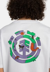 Camiseta blanca con un gráfico colorido de un círculo que contiene elementos terrestres en verde y púrpura, mostrando varios insectos e ilustraciones estilizadas.