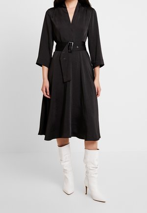 Woman wearing a black midi dress with three-quarter sleeves, a wide belt at the waist, and white pointed-toe heeled boots.