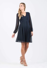 Marineblaue Chiffonkleid mit langen Ärmeln, V-Ausschnitt und Empire-Taillierung. Strukturierte Falten verleihen Volumen, kombiniert mit goldenen High-Heel-Sandalen.