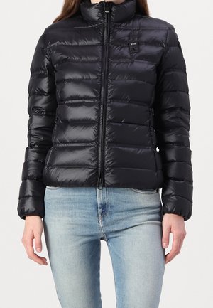 Vrouw draagt een zwarte gewatteerde donsjas met een ritssluiting aan de voorkant en lichtblauwe spijkerbroek, staand tegen een effen achtergrond.