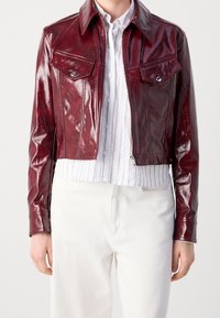 Krótkia, błyszcząca bordowa skórzana kurtka z dwoma kieszeniami na piersi, ostrym kołnierzem i zamkiem błyskawicznym z przodu. Nosi się ją na białej koszuli w paski.