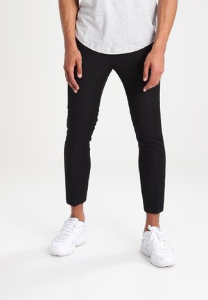 Mann trägt schmale schwarze Hose, weiße Sneakers und ein hellgraues T-Shirt und steht vor einem einfarbigen weißen Hintergrund.