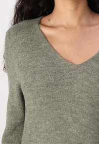 Pull vert olive avec un décolleté en V profond, fabriqué en tissu doux et texturé, doté de manches longues et d'une silhouette ajustée.