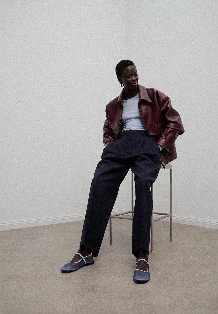 Bordowa skórzana kurtka, jasnoniebieski top, granatowe spodnie z wysokim stanem i niebieskie buty z detalami pasa; siedzi na metalowym stołku w minimalistycznym otoczeniu.