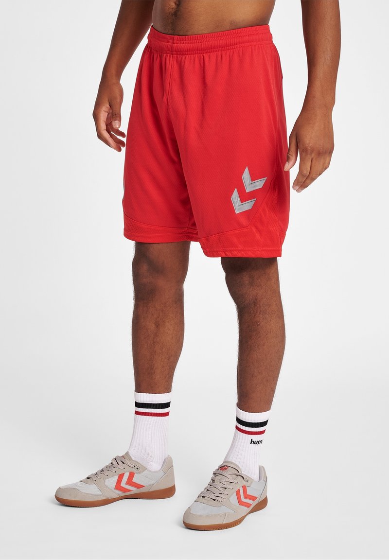 Homme portant un short de sport rouge avec des logos en forme de flèche blancs, des chaussettes blanches avec des rayures noires et rouges, et des baskets beiges avec des accents rouges.