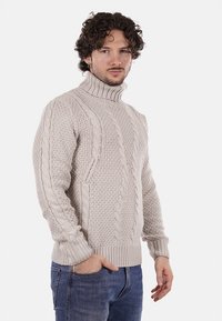Maglione beige chiaro a coste concollo alto, caratterizzato da motivi testurizzati e polsini a coste. Abbinato a jeans in denim blu.