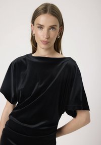 Černý sametový top s širokým lodičkovým výstřihem, draped krátkými rukávy a jemnou texturou, zdůrazňující uvolněnou, plynulou siluetu.