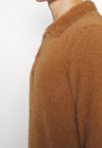 Brun, fluffig stickad cardigan med krage, knappstängning och mjuk textur, visad från sidan mot en ljus bakgrund.