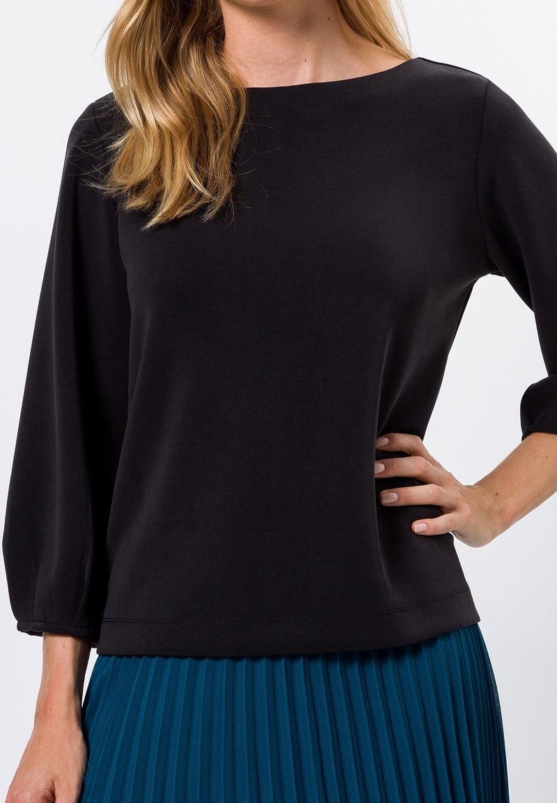 Maglione nero con scollatura ampia e maniche a tre quarti, realizzato in un tessuto morbido. Il design è semplice e informale, ideale per essere abbinato a una gonna plissettata color turchese.