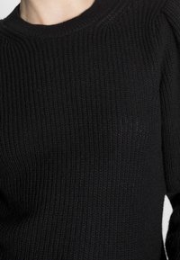 Schwarzer gerippter Strickpullover mit rundem Ausschnitt. Die Textur ist dick und strukturiert und weist subtile Variationen im Strickmuster auf.