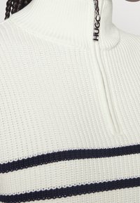 Suéter de punto blanco con canalé y cuello alto, rayas azul marino en el pecho y un tirador de cremallera etiquetado como "HUGO."