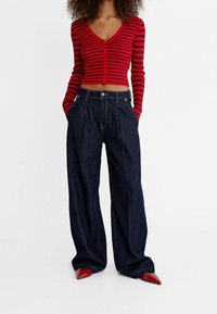 Rood en zwart gestreepte cropped cardigan met V-hals en knoopsluiting, gecombineerd met hoge taille donkere denim wijde broek en rode puntige schoenen.