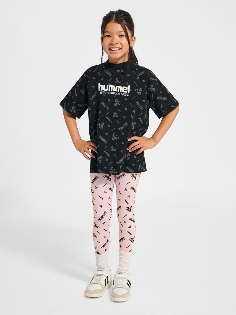 Lachend meisje staat met de handen in de zij, draagt een zwart Hummel Performance-shirt en roze leggings met logo's, witte sokken en sneakers.