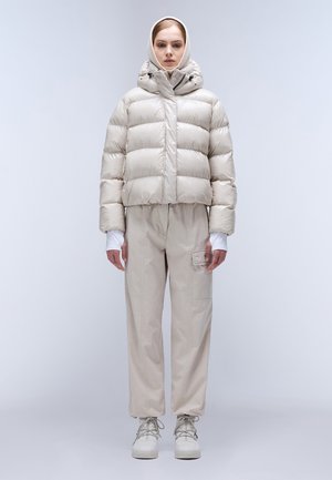 Zachte, lichte, korte jas in glanzend beige, gecombineerd met losse beige broek met een zijzak en witte sneakers.