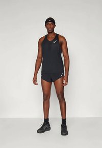 Un atleta masculino se encuentra de pie, vistiendo una camiseta sin mangas negra de Nike, pantalones cortos, una cinta para la cabeza, zapatillas negras y calcetines, todo ello contra un fondo gris liso.