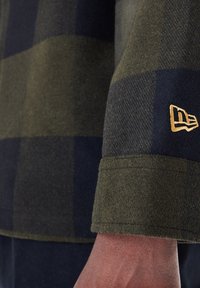 Groen en marineblauw geruit flanellen shirtstof met een gladde textuur. Heeft een gouden logo-detail op de manchet. Manchetmouw ontwerp.
