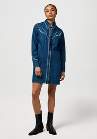 Abito camicia in denim blu scuro con dettagli di cuciture bianche, chiusura anteriore con bottoni, maniche lunghe e colletto. Indossato con stivali neri alla caviglia.
