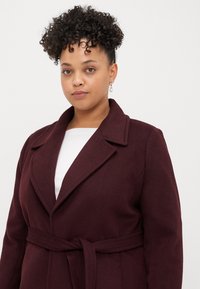 Μπορντώ παλτό με ζώνη και με σαρκώδη γιακά, φτιαγμένο από μαλακό ύφασμα. Διαθέτει λεία υφή και δομημένο σχέδιο.