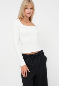 Witte blouse met lange mouwen en een vierkante halslijn, gecombineerd met zwarte broek met drawstring. De stof lijkt zacht met een gladde textuur.
