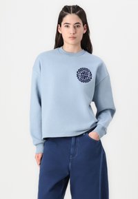 Hellblaues Sweatshirt mit einem runden navyfarbenen Grafiklogo auf der Brust. Verfügt über einen Rundhalsausschnitt und einen lässigen Schnitt. Kombiniert mit dunkelblauen Hosen.