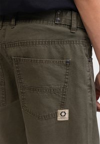 Pantalons en coton vert olive avec une surface texturée, des poches latérales et des coutures visibles. Une étiquette en tissu avec un logo est présente.