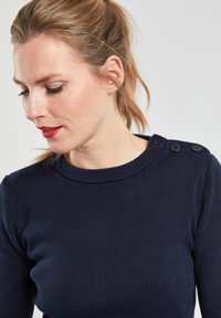 Pull en maille côtelée bleu marine avec un col rond, présentant des détails de boutons sur l'épaule gauche. Texture douce avec un design ajusté.